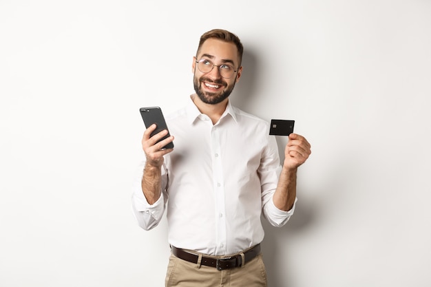 Business and online payment. Image of handsome man thinking while holding credit card and smartphone, standing against white background