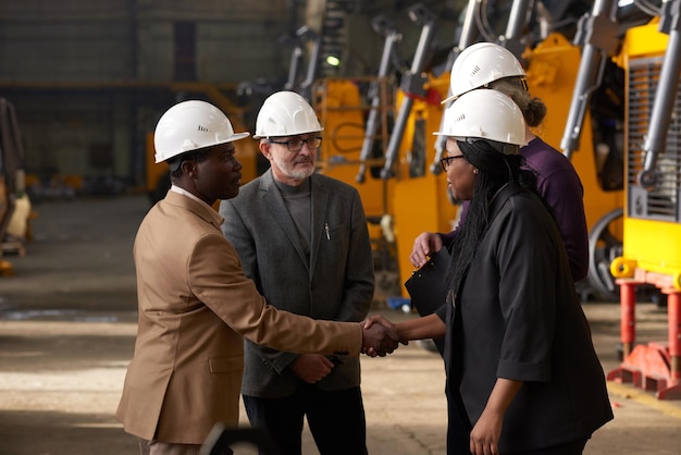 Business partners in work helmets shaking hands to conclude a deal during meeting on construction site with machines in the background