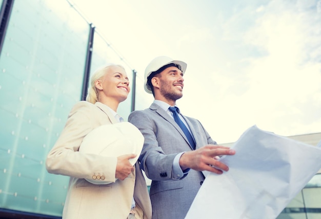 business, partnership, architecture and people concept - smiling businessman and businesswoman with blueprint and helmets on city street