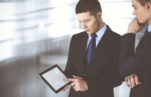 Business people are using a tablet computer, while standing in a modern office. Unknown businessman with a colleague at workplace. Teamwork and partnership concept.