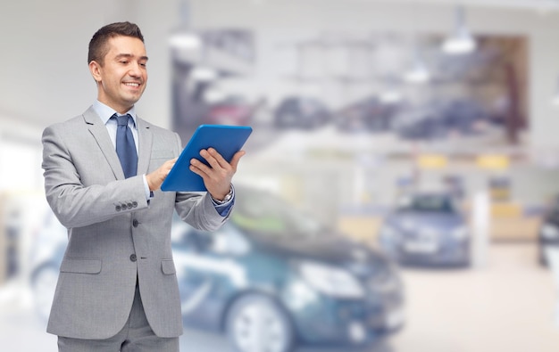 business, people, car sale and technology concept - happy smiling businessman in suit holding tablet pc computer over auto show or salon background