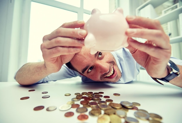 business, people, finances and money saving concept - happy businessman shaking piggy bank and getting coins at office