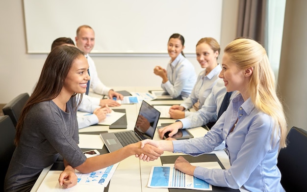 business, people, gesture and partnership concept - smiling business team with laptop computer and papers shaking hands in office