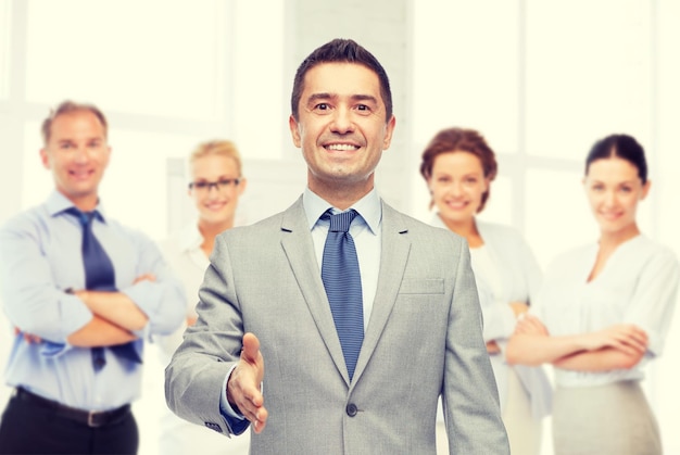 business, people, gesture, partnership and greeting concept - happy smiling businessman in suit with team over office room background shaking hand