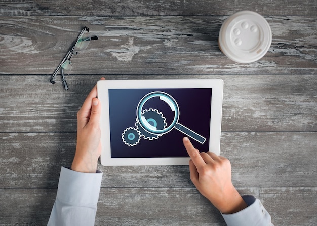 business, people, setting and technology concept - close up of hands pointing finger to tablet pc computer screen showing magnifier and cogwheel, coffee cup and eyeglasses on table