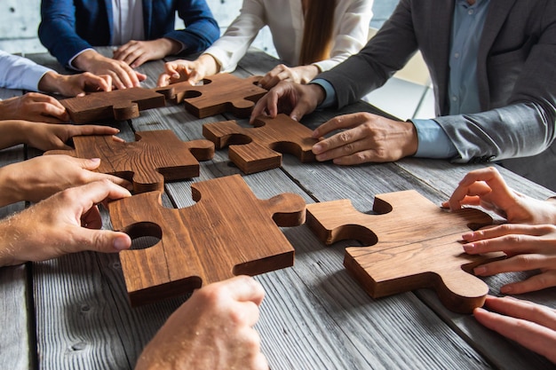 Business people team sitting around meeting table and assembling wooden jigsaw puzzle pieces unity cooperation ideas concept