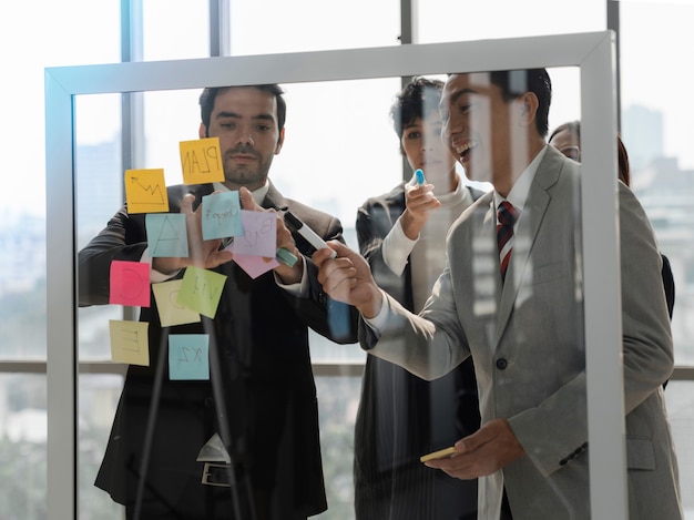 Business people behind transparent glass wall brainstorming and discussing at office and use post it sticky paper notes to share idea, team planing or new idea thinking concept