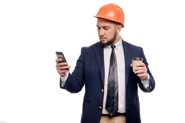 Business portrait of surprised contractor and developer, talking about phone. Businessman in hard hat with cup of coffee standing over white background. News and coffee brake concept