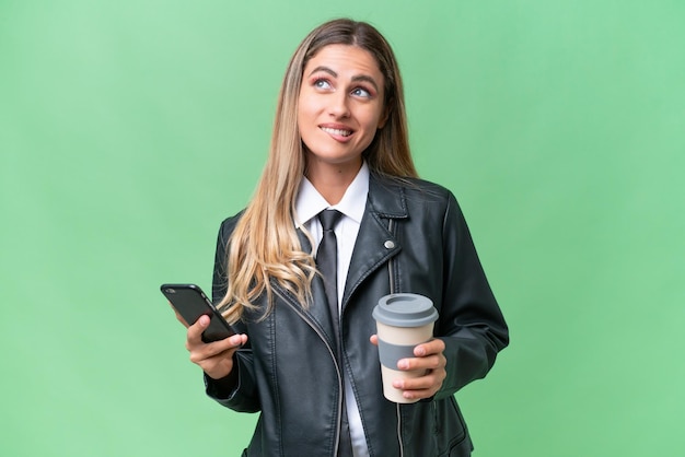 Business pretty Uruguayan woman wearing a biker jacket over isolated background holding coffee to take away and a mobile while thinking something