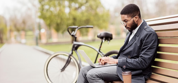 A business professional black man uses his laptop on a park bench beside his bike the scene is calm