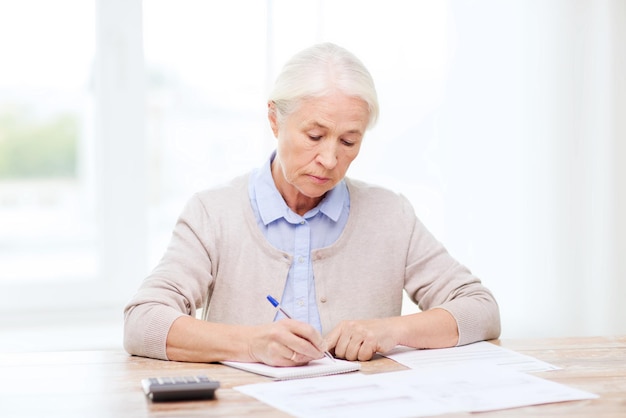 business, savings, annuity insurance, age and people concept - senior woman with papers or bills and calculator writing at home