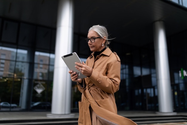 Business senior adult woman with a tablet walking down the street next to the office building year