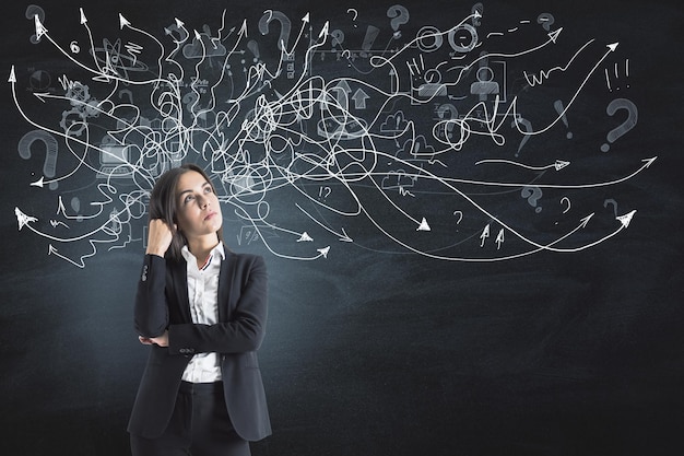 Business solution and brainstorming concept with pensive young woman in black suit looking up on dark wall background with hand drawn white chaotical arrows