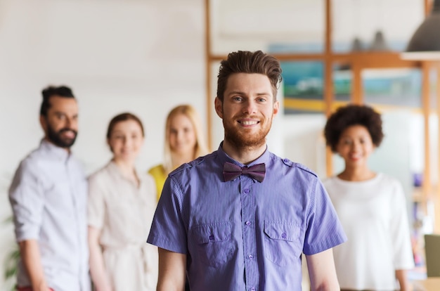 business, startup, people and teamwork concept - happy young man with beard and bow tie over creative team in office