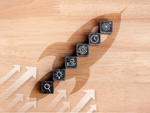 Business startup with growth success process for leadership concept. The business strategy icons on the black cube blocks as steps on rocket symbol with rising up arrows on wooden background.