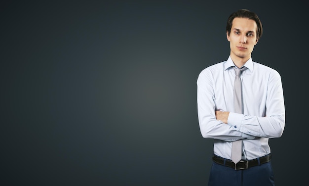 Business success concept with young smiling businessman folding his hands in white shirt and dark blue trousers on abstract black blank background with place for your logo or text Mock up