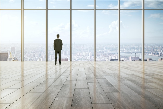 Business success leadership and inspiration concept with businessman back view looking on city skyline through big window in empty spacious hall with wooden floor