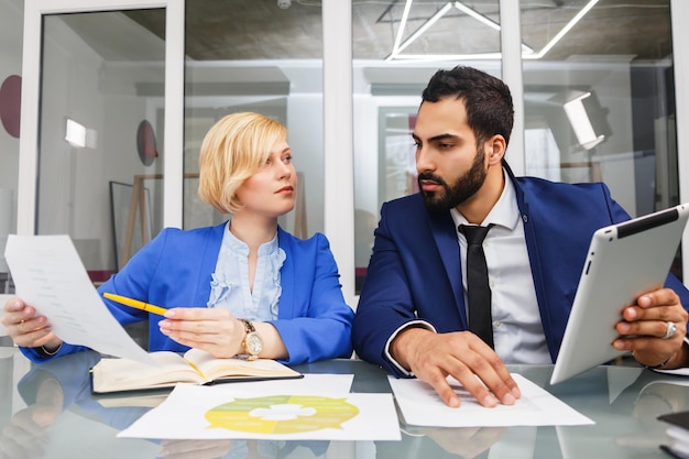 Business teammates office workers man and woman have discussion in glass room sitting on table with documents and tablet computer