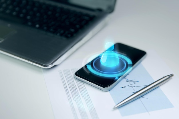 business, technology and communication concept - close up of smartphone with contact icon hologram, laptop computer and chart with pen on office table