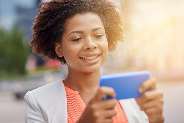 business, technology, communication and people concept - young smiling african american businesswoman with smartphone reading message on city street