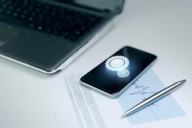 business and technology concept - close up of smartphone with cogwheel projection on screen, laptop computer and chart with pen on office table