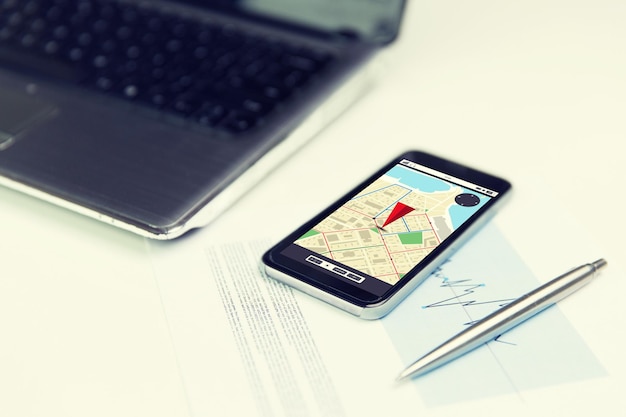 business, technology, navigation and location concept - close up of smartphone with gps navigator map on screen, laptop computer and pen on office table