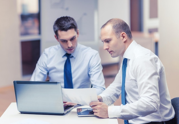 business, technology and office concept - two businessmen with laptop, tablet pc computer and papers having discussion in office