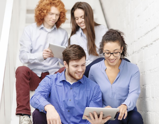 business, technology and startup concept - smiling creative team with tablet pc computer sitting on staircase