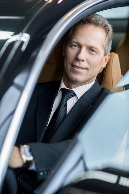 Business trip. Confident senior businessman sitting in car and smiling at camera