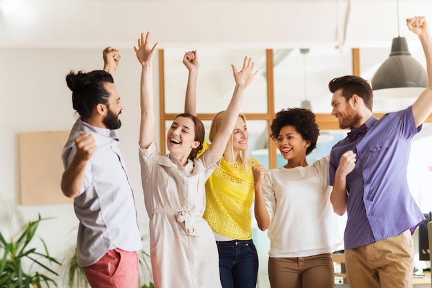 business, triumph, gesture, people and teamwork concept - happy international creative team raising hands up and celebrating victory in office