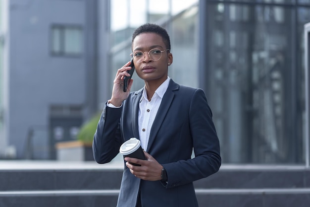 Business woman in business attire talking on the phone, African American woman on a break with a cup of hot drink walks in the business district near the office