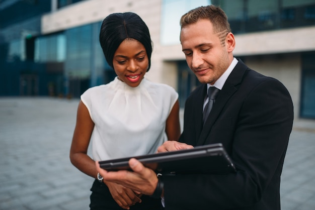 Business woman and businessman looks on laptop screen, outdoors meeting of partners