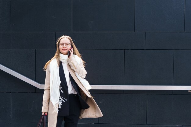 Business woman of caucasian ethnicity with glasses talking on the phone near the business center in ...