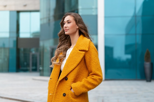 Business woman dressed yellow coat standing outdoors corporative building background Caucasian female business person on city street near office building with windows Stylish businesswoman