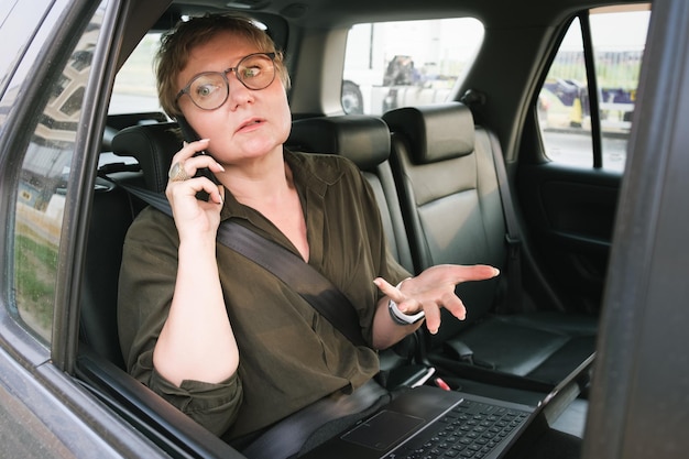 Business woman in glasses is talking on the phone while sitting in the back seat of a car