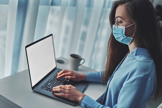 Business woman in glasses and medical protective mask online working from home at the laptop during self isolation and quarantine.