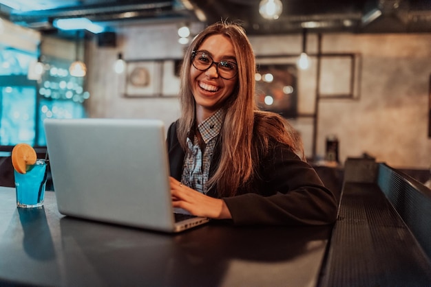 Business woman in glasses sitting in a modern cafe and working on her laptop while drinking a cocktail Selective focus