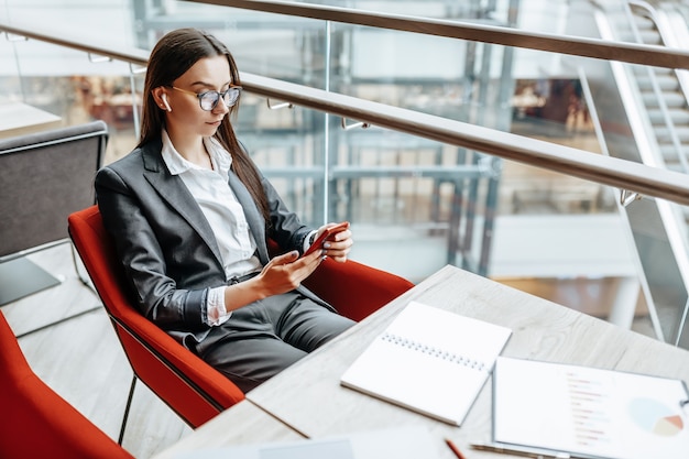 Business woman in glasses at the workplace uses the phone and sits at the table. Manager in the office.