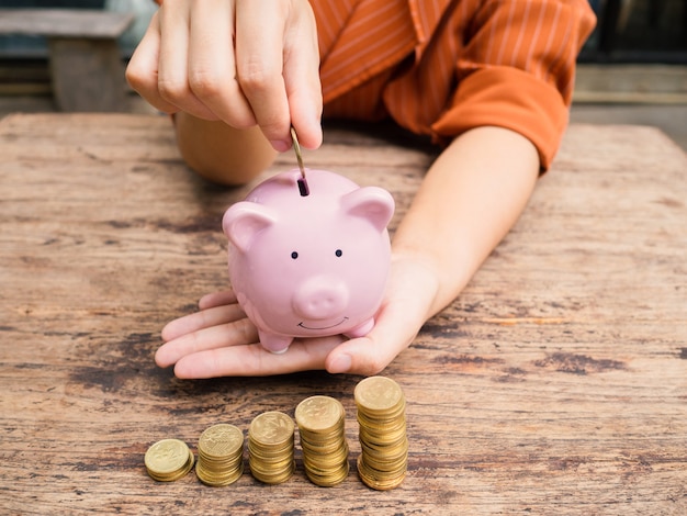 Business woman hand putting coin into  Pink piggy bank with 
gold coins pile growth graph, Saving money for future investment plan and retirement fund concept.