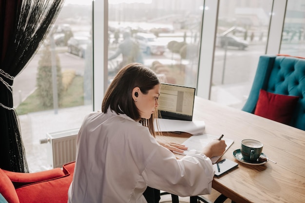 A business woman is sitting at a table in a cafe with documents and working on a laptop