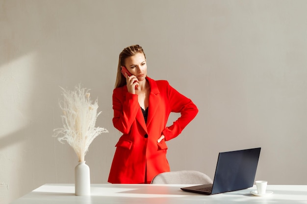 Business woman is talking on the phone in her office laptop in the table white wall background