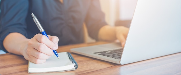 Business woman is writing on a notebook with a pen and using a laptop to work in the office.