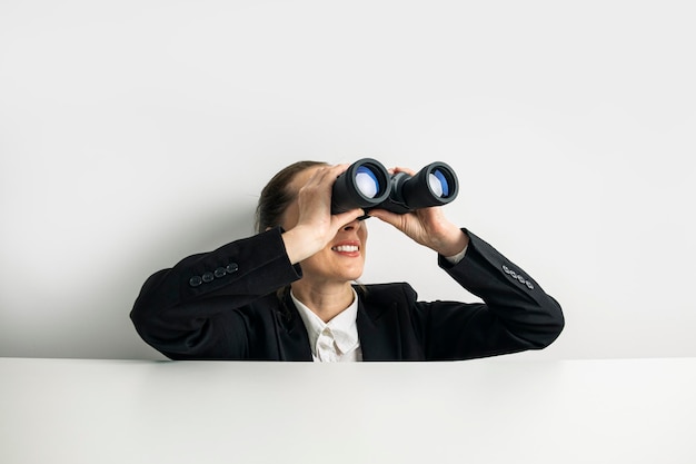 Business woman looking through binoculars peeking out from behind white table