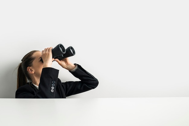 Business woman looking through binoculars peeking out from behind white table