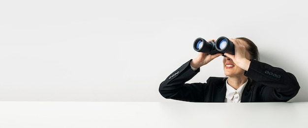 Business woman looking through binoculars peeking out from behind white table