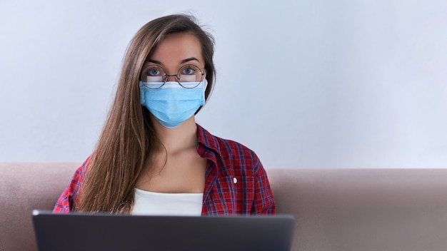 Business woman in round glasses wears medical protective mask working from home at the computer during self-isolation and quarantine.