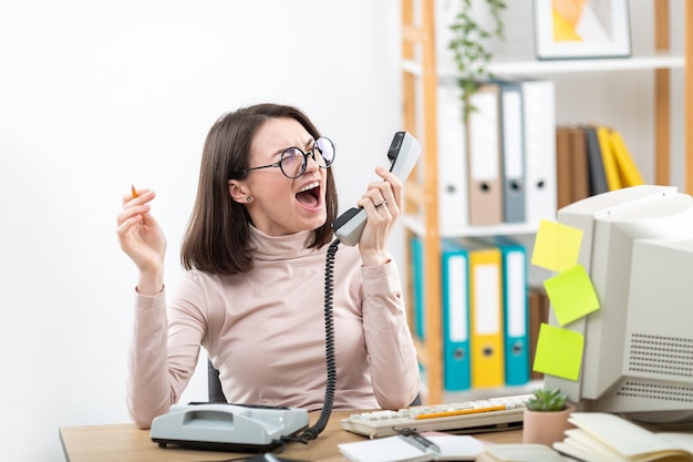 Business woman screaming into telephone receiver at work desk