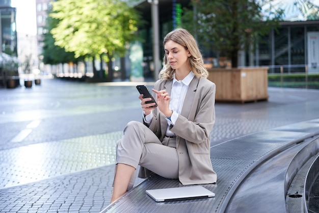 Business woman sits in city centre and works on mobile phone she has her laptop and wears working co