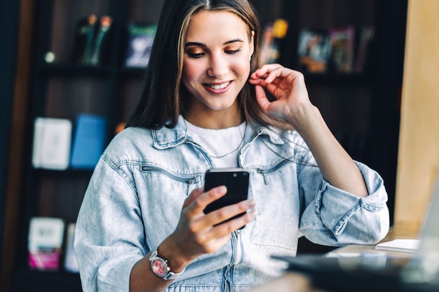 Business woman sits at table, holds smartphone in her hands, writes messages, conducts business correspondence.