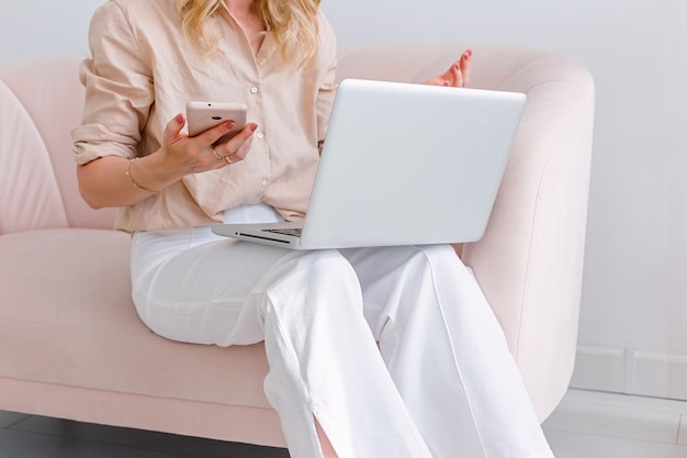 A business woman sits with a gray open laptop on her knees holds a pink smartphone